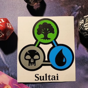 May include: A sticker featuring a green, blue and grey symbol representing the Sultai colour combination in the Magic: The Gathering card game. The symbol is a three-part design with a tree, a skull and a teardrop.