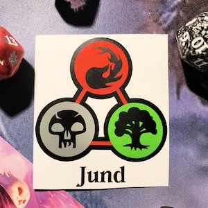 May include: A white sticker with a black, red, and green symbol. The symbol is a triangle with three circles inside. The circles are a black skull, a red and black flame, and a green tree. The word "Jund" is printed below the symbol.