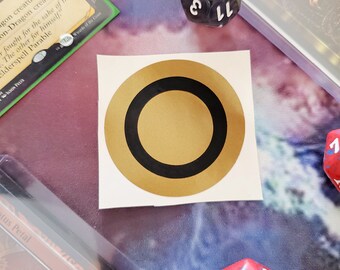 May include: A gold circle sticker with a black ring around the outside. The sticker is on a white background.