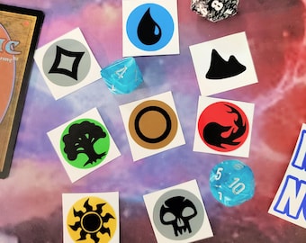 May include: A set of ten Magic: The Gathering mana symbols on white square stickers. The symbols are black, white, blue, red, green, and gold. The symbols are arranged in a grid pattern. There are two dice in the image, one black and white and one blue.