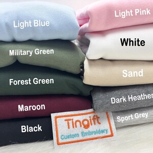 May include: A stack of folded sweatshirts in various colors, including light blue, military green, forest green, maroon, and black. Other colors are light pink, white, sand, dark heather, and sport grey. A label reads "Tingift Custom Embroidery."