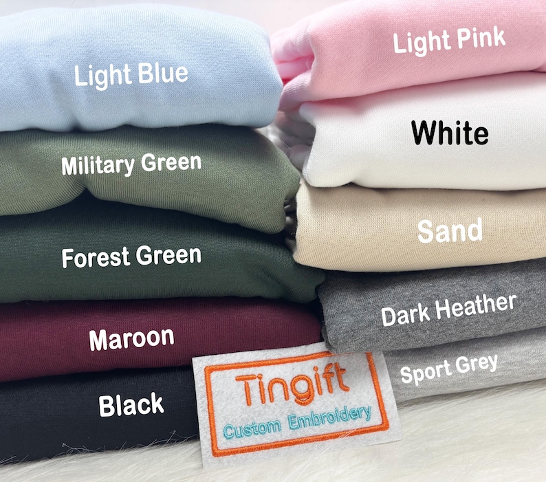 May include: A stack of folded sweatshirts in various colors, including light blue, military green, forest green, maroon, and black. Other colors are light pink, white, sand, dark heather, and sport grey. A label reads "Tingift Custom Embroidery."
