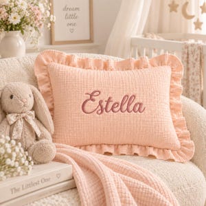 Peut inclure: Un coussin rectangulaire en mousseline couleur pêche avec un bord à volants et le prénom Estella brodé en écriture cursive rose foncé. Le coussin présente une texture en tissu froissé doux et est placé au centre de l'image.