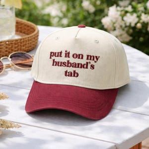 Può includere: Cappellino da baseball color crema con visiera e bottone superiore bordeaux. La frase "put it on my husband's tab" è ricamata in filo bordeaux sul davanti. Il cappello è su un tavolo bianco all'aperto.