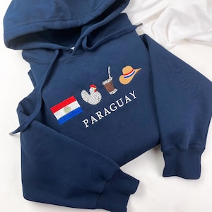 Könnte beinhalten: Marineblauer Kapuzenpullover mit gestickter Paraguay-Flagge, Huhn, Getränk und Hut. Das Wort "PARAGUAY" ist weiß gestickt. Der Pullover hat eine Kapuze mit Kordelzug und gerippte Bündchen.