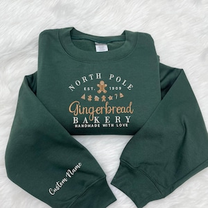 Może przedstawiać: Ciemnozielona bluza z napisem "NORTH POLE EST. 1909 Gingerbread BAKERY HANDMADE WITH LOVE" w kolorze złotym i białym. Lewy rękaw ma napis "Custom Name" w kolorze białym.