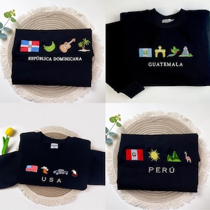 Op de afbeelding: Vier zwarte sweatshirts met geborduurde ontwerpen. Eén heeft de vlag van de Dominicaanse Republiek, bananen, een gitaar en palmbomen. Een ander toont de Guatemalteekse vlag, een gebouw en een vogel. De derde heeft de Amerikaanse vlag, een vogel, een auto en een adelaar. De laatste toont de Peruaanse vlag, zon, bergen en een lama.
