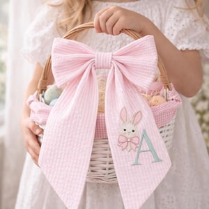 May include: A white wicker Easter basket with a pink gingham lining and a large pink bow. A pink fabric panel with a bunny and the letter "A" is attached to the basket. The basket is filled with pastel-coloured eggs.