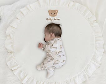 May include: A baby lies on a white, ruffled, circular play mat. The mat features a small embroidered bear and the text "Baby Name." The baby wears a patterned onesie. A stuffed animal is visible in the background.