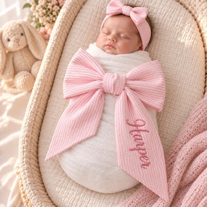 May include: A newborn baby wrapped in a white swaddle blanket featuring a large, pink and white striped bow with the name Harper embroidered in pink script. The baby wears a matching pink and white striped headband with a small bow on top.