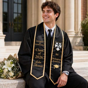 May include: A graduate in a black gown with a gold-edged sash from the University of Pennsylvania. The sash reads "Class of 2026", "John Thomas", and "BS Biological Sciences". The graduate smiles, holding a bouquet of white flowers.