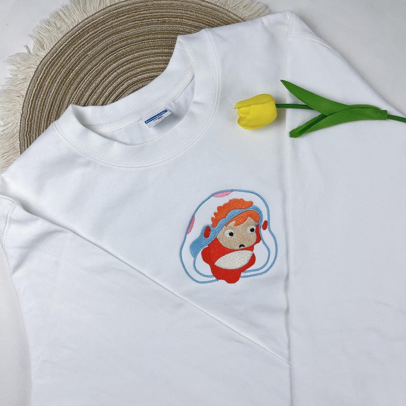 May include: White sweatshirt featuring an embroidered cartoon character in red, orange, and blue. The design is centered on the front of the long-sleeved crew neck shirt. A yellow tulip and a woven mat are in the background.