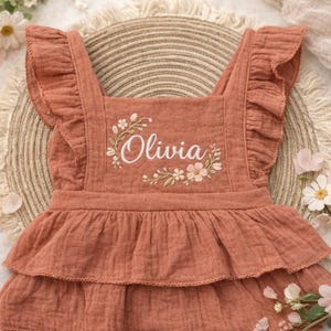 May include: A terracotta-colored baby romper with ruffled straps and a tiered skirt. The name "Olivia" is embroidered in white script, surrounded by floral accents. The romper is made of a soft, textured fabric, perfect for a baby.