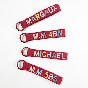 Puede incluir: Cuatro llaveros de tela roja con texto bordado. Los llaveros tienen un anillo de metal y presentan los nombres MARGAUX, MICHAEL, e iniciales con números y letras en varios colores.
