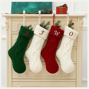May include: Four cable-knit Christmas stockings in green, white, and red, each with a different initial embroidered on the cuff. The stockings are suspended from white hooks, with greenery and berries inside.