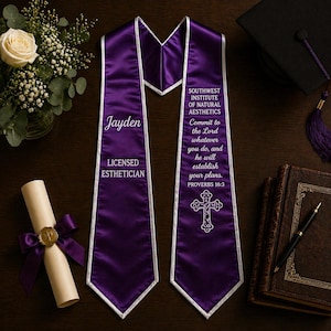 May include: A purple graduation sash with white edging and text. The sash displays "Jayden" and "Licensed Esthetician" on one side, and on the other, "Southwest Institute of Natural Aesthetics" and a bible verse. A diploma, flowers, and a graduation cap are also visible.