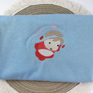 May include: A light blue sweatshirt featuring an embroidered cartoon character in a red outfit and a clear bubble. The character has brown hair and a worried expression. The embroidery is detailed and the sweatshirt is folded.