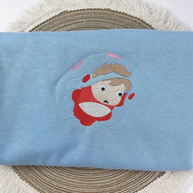 May include: A light blue sweatshirt featuring an embroidered cartoon character in a red outfit and a clear bubble. The character has brown hair and a worried expression. The embroidery is detailed and the sweatshirt is folded.
