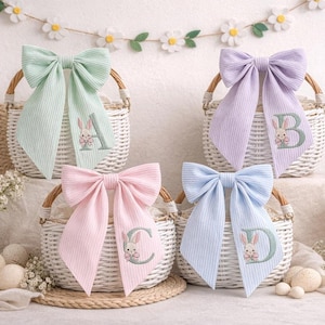 May include: Four Easter baskets with large, striped bows in pastel shades: mint green, lavender, pink, and light blue. Each bow features a letter and a bunny embroidery. The baskets are white with wooden handles.