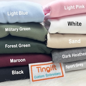 May include: A stack of folded sweatshirts in various colors, including light blue, military green, forest green, maroon, and black. Other colors are light pink, white, sand, dark heather, and sport grey. The label reads "Tingift Custom Embroidery."