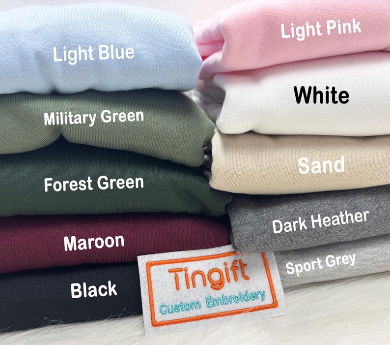May include: A stack of folded sweatshirts in various colors, including light blue, military green, forest green, maroon, and black. Other colors are light pink, white, sand, dark heather, and sport grey. The label reads "Tingift Custom Embroidery."