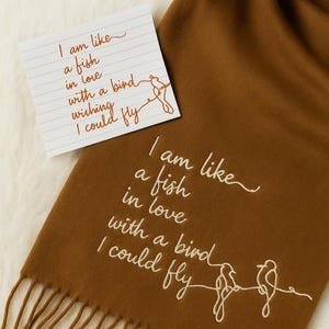 May include: A brown scarf with white embroidered text and bird illustrations. The text reads "I am like a fish in love with a bird wishing I could fly." A small card with the same text is also present.