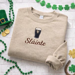 May include: A tan sweatshirt with a black and white embroidered beer glass and the word "Sl&aacute;inte" in brown. The sweatshirt is surrounded by St. Patrick's Day decorations, including a green hat, gold coins, and shamrocks.