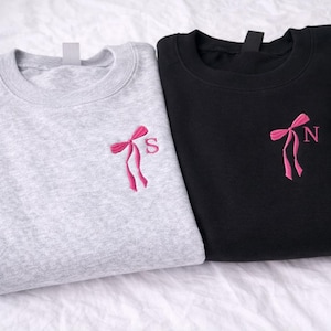 Könnte beinhalten: Zwei Sweatshirts, eines hellgrau und eines schwarz, werden gezeigt. Jedes hat eine rosa gestickte Schleife mit einem einzelnen Buchstaben. Das hellgraue Sweatshirt hat den Buchstaben "S", während das schwarze den Buchstaben "N" hat. Beide sind aus weichem Material.