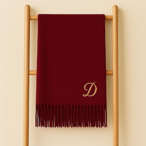 Könnte beinhalten: Ein weinroter Schal mit einem goldgestickten Buchstaben "D" und Fransen. Der Schal ist auf einem hölzernen Kleiderständer drapiert, der seine Textur und sein Design zeigt. Der Schal ist einfarbig und scheint aus einem weichen Material zu sein.