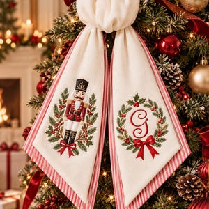 May include: A cream-coloured fabric tree topper bow with two long hanging tails. One tail features an embroidered nutcracker surrounded by a holly wreath, while the other displays a red embroidered letter G inside a holly wreath with a red bow.