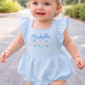 May include: A baby's light blue romper and headband set. The romper has a square neckline, frilled sleeves, and the name "Michelle" embroidered in cursive. Floral embroidery decorates the bodice. The outfit is made of a soft, textured fabric.