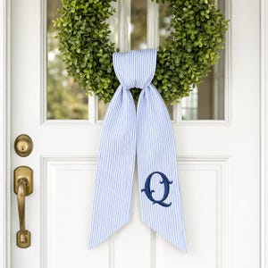 Puede incluir: Una corona verde cuelga de una puerta blanca, adornada con una cinta a rayas azules y blancas. La cinta presenta una letra "Q" bordada en azul marino. La puerta tiene una manija y cerradura de latón.