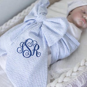 Può includere: Una coperta per neonati a righe blu e bianche con un grande fiocco. La coperta presenta un monogramma ricamato blu scuro. Il bambino indossa un cappello bianco e un body bianco.