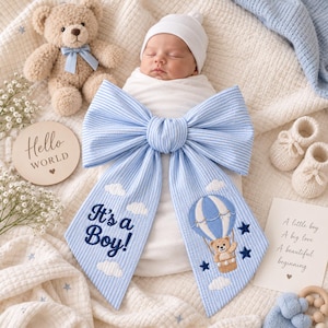 Può includere: Un neonato avvolto in una coperta bianca, con un berretto bianco, decorato con un grande fiocco a righe blu e bianche con il testo ricamato It's a Boy, una mongolfiera con un orsetto, nuvole e stelle.