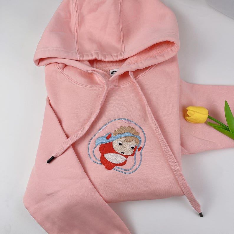 May include: A light pink hooded sweatshirt with a drawstring and a cartoon character embroidered on the front. The character is wearing a blue hat and red outfit. A yellow tulip is in the background.