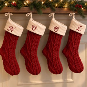 May include: Four red cable-knit Christmas stockings with white ribbed cuffs, each featuring a single embroidered letter in red thread. The letters N, O, E, and Z are stitched onto the cuffs. Each stocking includes a white hanging loop.