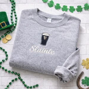 May include: A heather gray sweatshirt with an embroidered pint of stout and the word "Sl&aacute;inte" in white script. The sweatshirt is folded, with the sleeves cuffed. Green shamrock decorations and gold coins are visible.