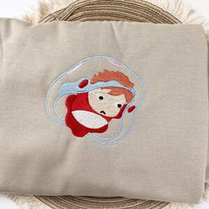 May include: A beige sweatshirt featuring an embroidered cartoon character in a red outfit and a blue bubble. The character has orange hair and a sad expression. The embroidery is detailed with blue, red, and black thread.
