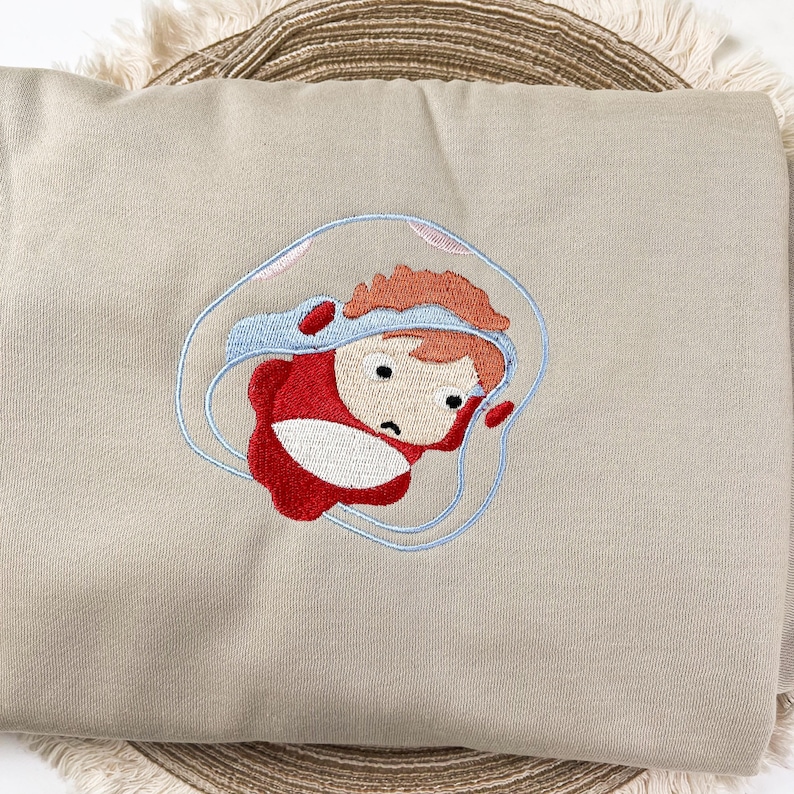 May include: A beige sweatshirt featuring an embroidered cartoon character in a red outfit and a blue bubble. The character has orange hair and a sad expression. The embroidery is detailed with blue, red, and black thread.