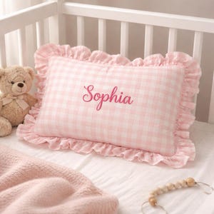May include: Pink gingham pillow with ruffled edges, embroidered with the name "Sophia" in a cursive font. The pillow is placed in a white crib with a teddy bear and a pink blanket.