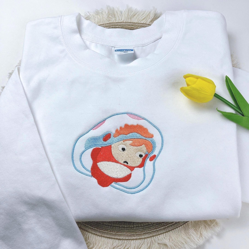 May include: White sweatshirt with an embroidered cartoon character in red, orange, and blue. The design features a character inside a blue and white shell-like shape. A yellow tulip is placed on the right side of the sweatshirt.