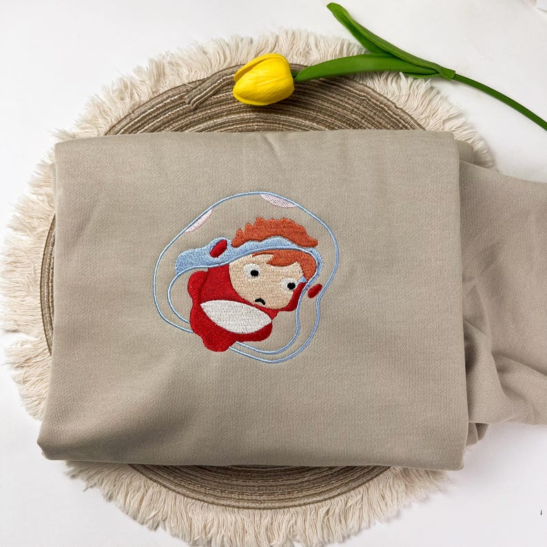 May include: A beige sweatshirt with an embroidered cartoon character in a red outfit and blue bubble. The character has red hair and a sad expression. The sweatshirt is folded on a round, woven mat.