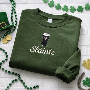 May include: Green sweatshirt with an embroidered pint of stout and the word "Sl&aacute;inte" in white script. The sweatshirt is surrounded by St. Patrick's Day decorations, including a green hat, gold coins, and shamrock garlands.