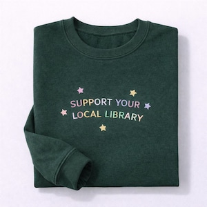 May include: Dark green sweatshirt featuring the text "SUPPORT YOUR LOCAL LIBRARY" in pastel colours. The text is surrounded by small star graphics. The sweatshirt has a crew neck and long sleeves.
