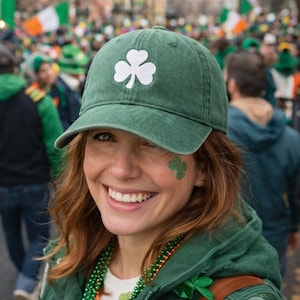 Può includere: Un berretto da baseball verde con un'emblema a trifoglio bianco. Il berretto è indossato da una persona con un trifoglio dipinto sulla guancia. La persona sorride e indossa perline verdi. Lo sfondo mostra una folla di persone.
