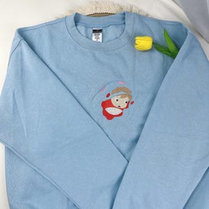 May include: A light blue sweatshirt with an embroidered design of a cartoon character in a red outfit. The sweatshirt has long sleeves and a crew neck. A yellow tulip is placed on the right side of the sweatshirt.