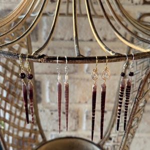 May include: A collection of handmade earrings displayed on a decorative metal stand. The earrings feature various designs, including dark red beads, wire-wrapped spirals, and elongated, striped elements in shades of brown and cream.