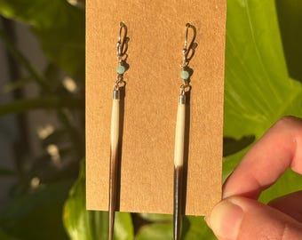 May include: A pair of handmade earrings featuring porcupine quills. Each earring has a silver-colored hook, a small turquoise bead, and a quill with a white and brown pattern. The earrings are displayed on a brown card.