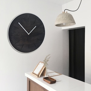 May include: A black clock with white hands and a silver frame hangs on a white wall above a wooden counter. A concrete lamp with a brass fixture hangs from the ceiling above the counter.