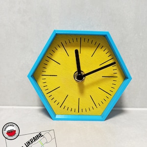 Könnte beinhalten: Eine gelbe sechseckige Uhr mit schwarzen Zeigern und einem blauen Rand. Die Uhr steht auf einer weißen Oberfläche. Ein Aufkleber mit dem Text "POLAND UKRAINE STAY TOGETHER" ist in der linken unteren Ecke des Bildes zu sehen.